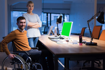 Handicapped young man with female  colleague working in office. Disabled businessman in the wheelchair works in the office at the computer while performing in co-working space. Looking at camera.