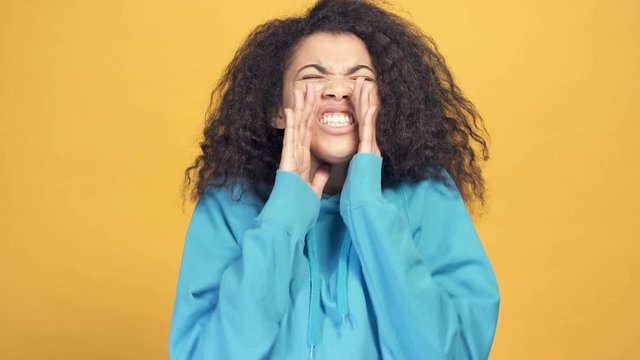 Close up portrait of young afro american woman shouting and looking at the camera. Yellow background.