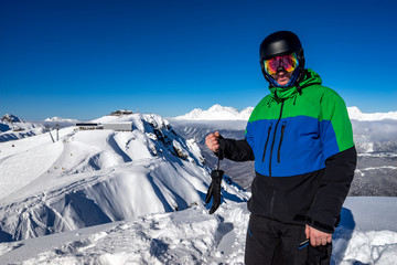 Fototapeta na wymiar Freerider snowboarder standing on steep slope of mountain peak, showing a sign is OK, on background of snowy mountains in ski resort