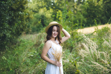 Naklejka na ściany i meble a cute and stylish girl in a white dress and long hair in the sunny summer park. A beautiful young girl in a white dress standing near the green tree