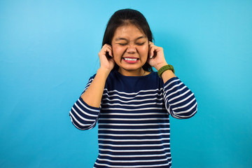 Portrait of Young beautiful asian women with blue isolated background, hears bothering noise, distressed frustrated woman plugs ears