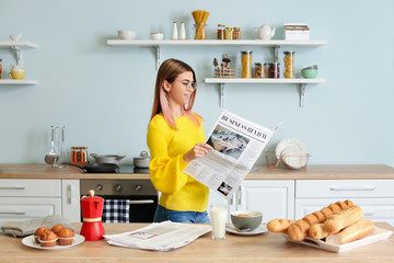 Beautiful young woman reading newspaper in kitchen