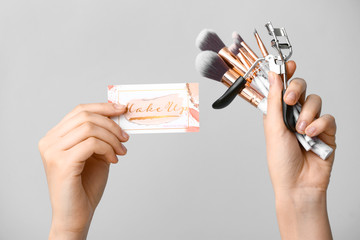 Female hands with business card of makeup artist and tools on grey background