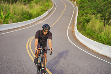 Obraz premium asian male riding a black bicycle on a road with a water bottle attached to it, smiling and, wearing bicycle helmet and goggles, with grey clouds, blue sky and tall forest trees in the background.