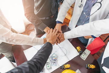 professionals putting hands together piling on top of one another, businessmen, doctors, architectures, engineer and colleagues, standing around table with money, glasses, and blueprint on the table