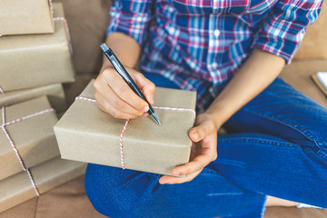 a person holding using a pen writing an address on the top of the wrapped up box of parcel with a string wrapped around it, sitting down on a couch next to multiple parcel piled on top on each other