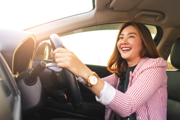 asian businesswoman driving a car holding a steering wheel with both hands while smiling joyfully, sitting in the driver seat with seatbelt on wearing suit and with sunset light shining in background