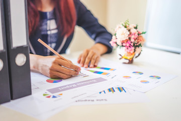 asian businesswoman looking down working and writing on a paper document using a pencil, working on data and statistics on graphs and charts, red long hair wearing a suit next to a cup of followers