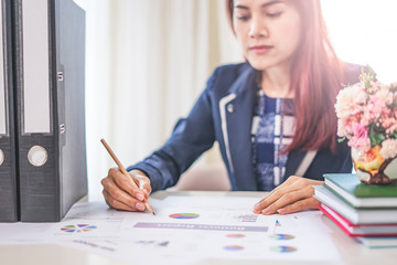 asian businesswoman looking down working and writing on a paper document using a pencil, working on data and statistics on graphs and charts, red long hair wearing a suit next to a cup of followers