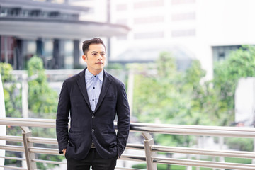 asian businessman standing and leaning on a railing, in a city urban area on a high architectural platform with tall buildings and green nature in the background, dressed formal wearing a black suit..