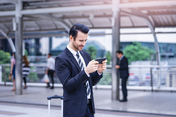 Caucasian businessman smiling joyfully playing on his smartphone, holding phone with both hands on landscape, wearing formally suit and tie with tall city skylines and metro station   background