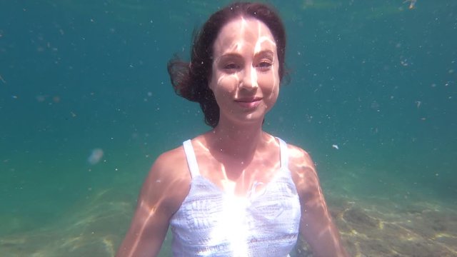 Young woman in a white dress expressing her emotions underwater, looking serious, smiling and touching her face elegantly