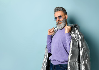 Gray-haired, aged man in purple pullover and sunglasses, silver colored down puffy jacket. Posing on blue background. Close up