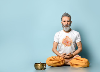 Yogi gray-haired man in dhoti clothes, holding rosary, sitting on floor in lotus pose on blue...