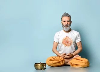 Fotobehang Yogi gray-haired man in dhoti clothes, holding rosary, sitting on floor in lotus pose on blue background. Singing bowl nearby © FAB.1