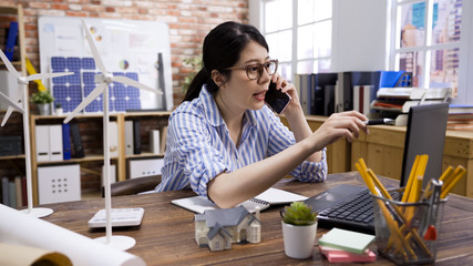 asian japanese female architect wearing eyeglasses working at desk on laptop computer. young lady employee talking on cellphone with coworker discussing on alternative energy architecture project.