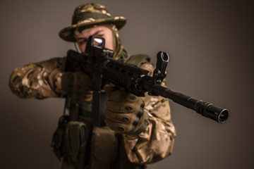Portrait of  soldier in military camouflage uniform protected with helmet, body armour, holding machine gun desaturated on a gray background.