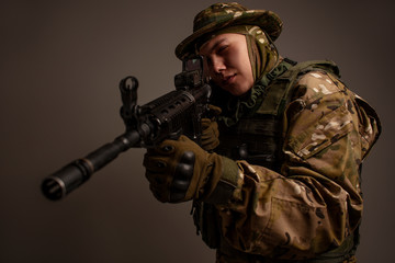 Portrait of  soldier in military camouflage uniform protected with helmet, body armour, holding machine gun desaturated on a gray background.