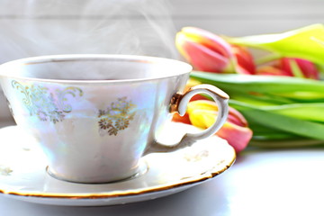 White porcelain pattern cup of hot black tea on dish with smoke on white desk with blurred Bouquet of flowers of red yellow blooming tulips with green leaf on backdrop. Selective focus