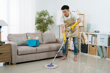 Asian japanese young man wearing apron cleaning floor at home. guy washing floor with mopping stick and bucket in living room of bright modern apartment. male household helping wife tidy house.