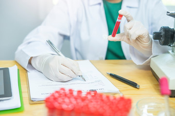 close up of female doctor or scientist wearing rubber gloves, goggles, mask and lab coat, holding test tube looking into the red fluid using a microscope, writing on a form documentation for analysis