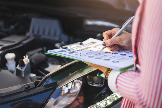 close up of asian female car inspector holding a pen clipboard checking on the engine with the paper work on the clipboard, with the car hood opened up revealing the car engine in the summer sun light