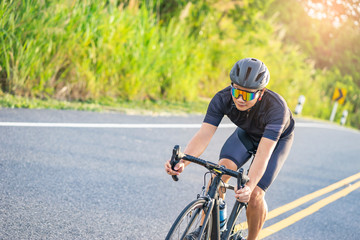 asian male riding on a black bicycle along the road, smiling and wearing bicycle gears, crash helmet and goggles, on a long winding road with forest trees and sunlight shining through the background.