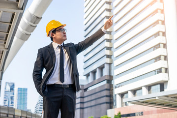 Asian male construction manager standing with hand in pocket and pointing a finger towards the tall building, formally wearing a suit and a hardhat for safety, within an urban city as the background