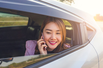 asian businesswoman smiling and looking out of the car window while sitting in the backseat, holding a smartphone against her ear and calling someone with evening sun set light shining in background