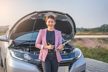 portrait of asian female car inspector holding a pen clipboard checking on the engine with the paper work on the clipboard with the car hood opened up revealing the car engine, leaning against the car