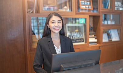 asian woman hotel receptionist standing behind a marble counter in front of a computer monitor...