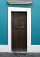 Simple brown wooden double doors with brown metal bars over the windows framed into a teal Turkish blue wall surrounded by white wooden trim on a grey brick sidewalk.
