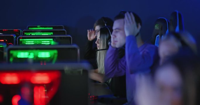 Team of young people plays in a computer club, an online game. Boys and girls are sitting in computer chairs with earphones and playing on the net. Computer club with modern technology.