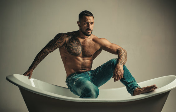 Sexual macho man in bath. Strong muscular tattoed man holding champagne bottle and posing in bathroom. Handsome bearded shirtless man in jeans with sexy body in bathroom.
