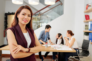 Pretty young confident businesswoman folding arms and smiling at camera when her team discussing project ideas