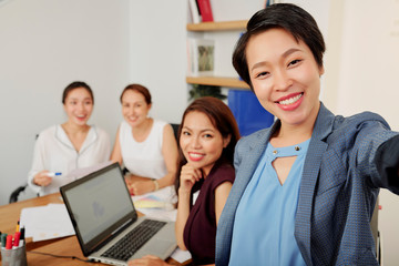 Cheerful young Vietnamese female entrepreneur taking photo with her smiling colleagues after successful meeting