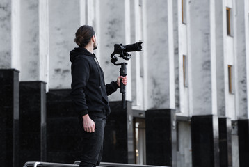 Fototapeta na wymiar Young Professional videographer holding professional camera on 3-axis gimbal stabilizer. Pro equipment helps to make high quality video without shaking. Cameraman wearing black hoodie making a videos.