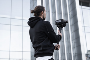 Young Professional videographer holding professional camera on 3-axis gimbal stabilizer. Pro equipment helps to make high quality video without shaking. Cameraman wearing white hoodie making a videos. © Volodymyr_sh