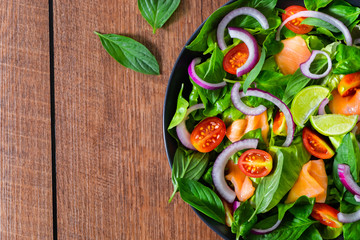 Top view from above of salmon salad with fresh tomato, onion, spinach, lettuce and basil in black bowl on wooden background. Homemade concept. Vegetables for tasty & healthy. Flat lay with copy space.