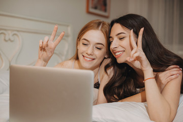 Two cheerful caucasian sisters lying in bed and talking with somebody using a laptop