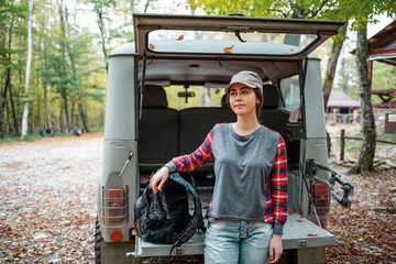 Fototapeta na wymiar A young Caucasian woman poses with a backpack against the open trunk of a jeep and looks away. In the background, trees and forest. Concept of car travel and tourism