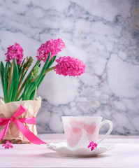 white cup with pink hearts with a vase of spring flowers on the table against the background of gray marble. Morning coffee, girl’s breakfast, spring mood