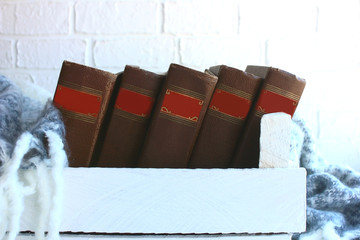 Vintage old books in a white wooden box with a mohair warm blanket on a background of a white brick wall. Collection of five antique books