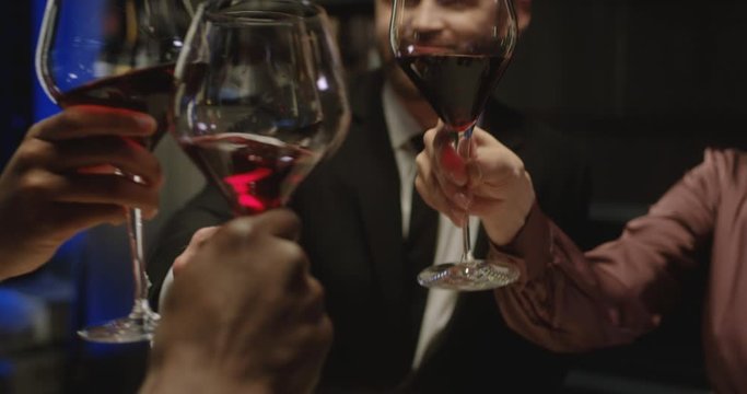 Close up of Caucasian and African American male and female people clashing with red wine in glasses and sipping at dinner in restaurant. Drinks in hands of mixed-races men and women.
