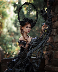 Obraz premium Portrait of amazing cute young woman with red hair with big black crown on her head and fabulous long dress put her hands on an old wrought iron gothic railing and looks at camera on blurry background