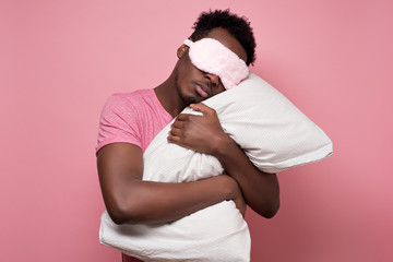 Young african american black man sleeping hugging a pillow wearing sleeping mask. Studio shot on...