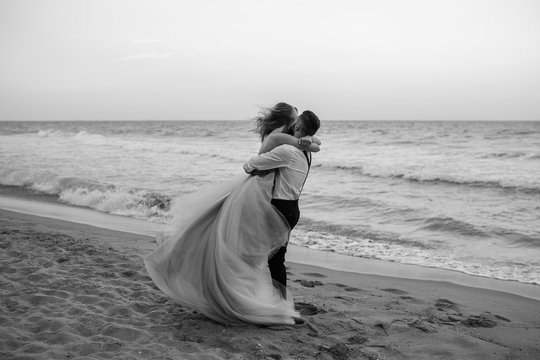 Bride and groom having fun on sand beach near the sea. Beach wedding concept. The groom holding bride in his arms. Love kissing. Black and white. Copy space.