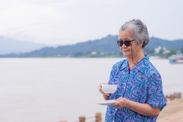 Fototapeta premium Elderly woman standing holding a white cup of coffee at side the river with nature background. Morning coffee. Space of text