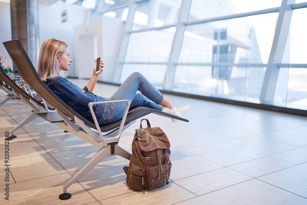 Wall mural travel and technology. pretty young woman using smartphone while sitting at airport terminal waiting