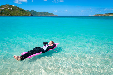 Relaxed man in suit floating on a bright pink pool raft in tropical blue waters
